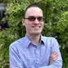 Smiling man with glasses wearing a light blue shirt standing outdoors in front of greenery.